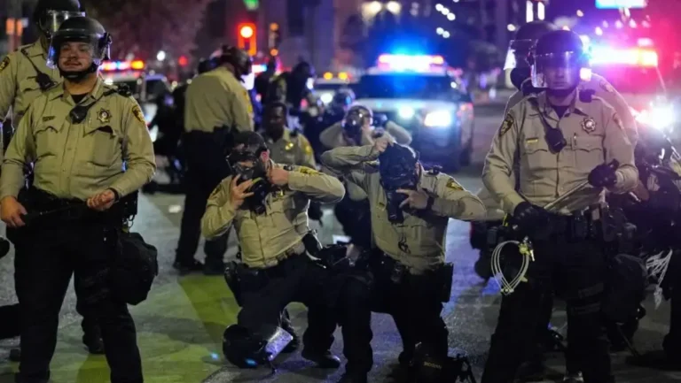 Agentes de la Policía Metropolitana de Las Vegas con luces de patrullas encendidas frente a un grupo de manifestantes durante una protesta nocturna en el centro de la ciudad; imagen relacionada con la demanda que acusa a la policía de uso excesivo de fuerza y disparos de gas pimienta contra manifestantes anti-ICE.
